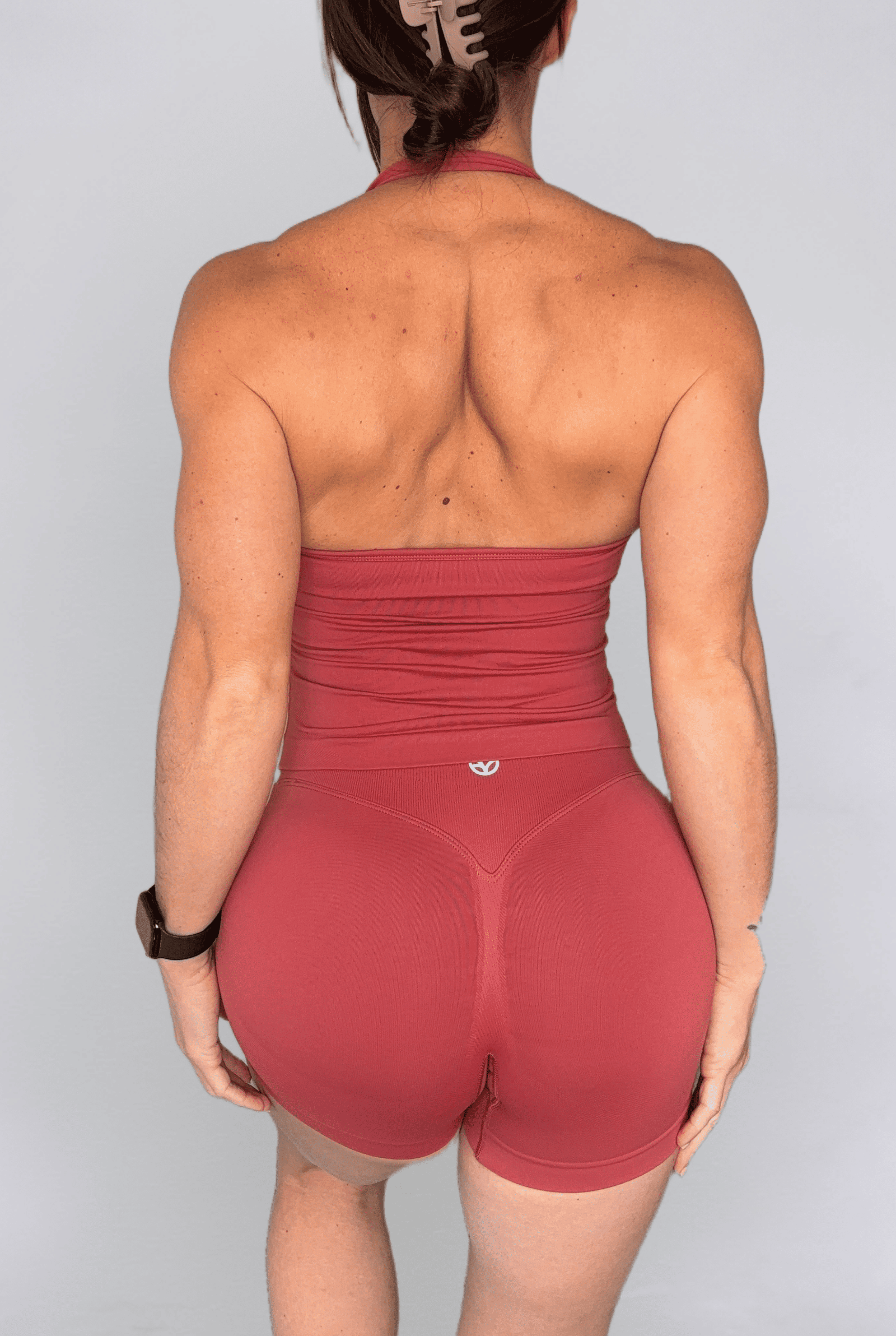 Rear view of a woman wearing a terracotta high-waisted athletic set consisting of a strapless tank top and matching high-waisted shorts, on a light gray background.
