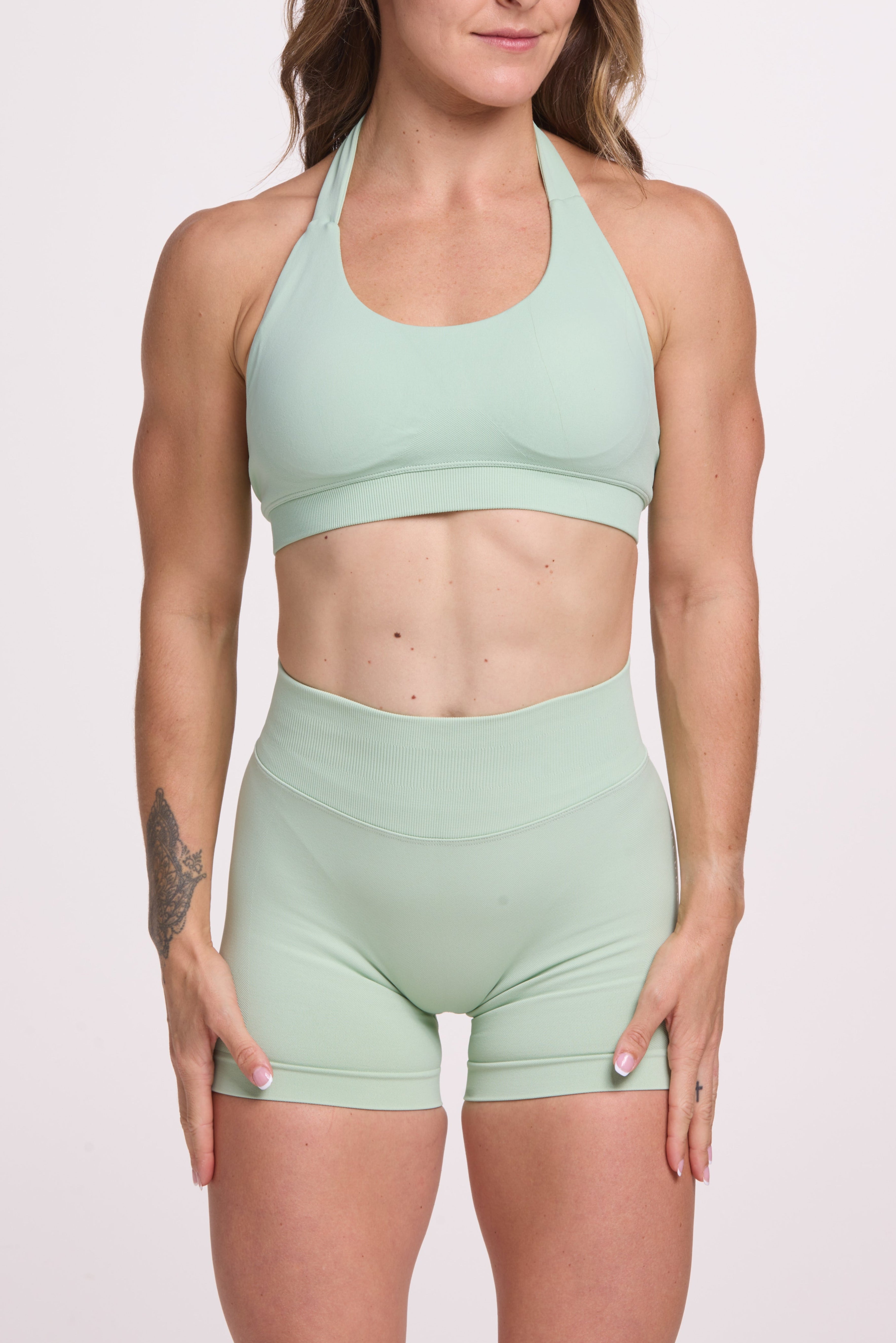 Seamless halter bra with a thick supportive fabric blend and a branded back strap, with a ribbed elastic hem and seamless gym shorts with booty contouring panels, invisible scrunch technology, sweetheart shaped back seams and thick supportive fabric in mist green