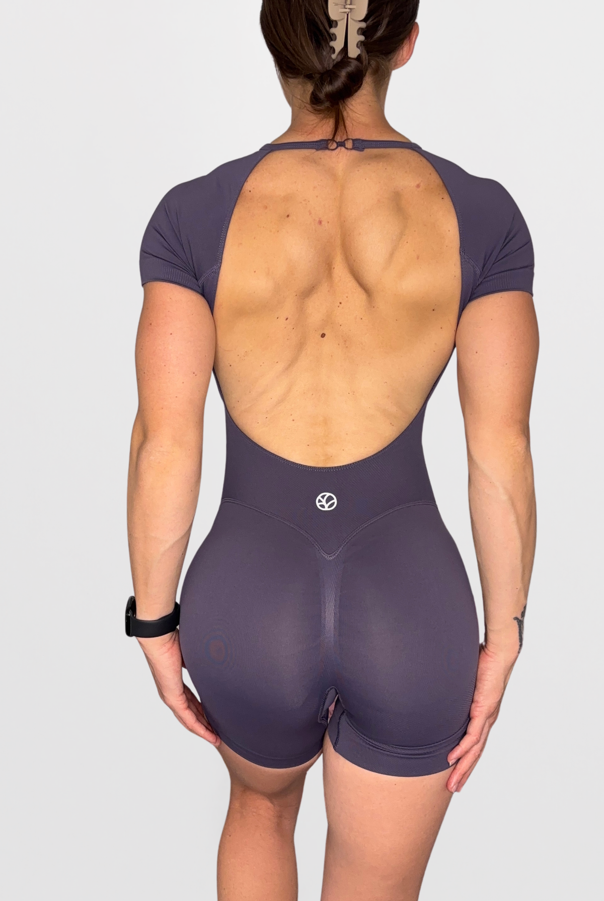 Rear view of a woman wearing a short-sleeve athletic bodysuit in Sweet Grape with a large open back, shown against a light grey studio background.