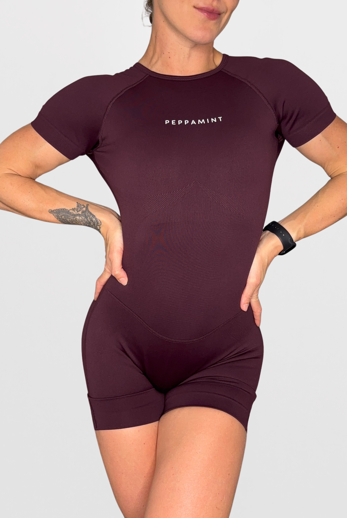 A woman wearing a short-sleeve athletic bodysuit in deep plum purple with 'PEPPAMINT' printed in white on the chest, set against a solid light grey background.