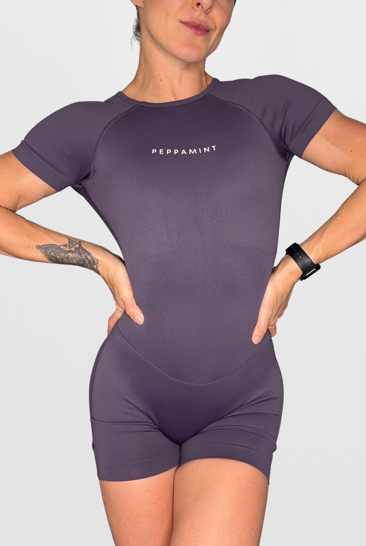 A woman wearing a short-sleeve athletic jumpsuit in a muted dusty purple colour, featuring the brand name 'PEPPAMINT' printed in white on the chest. The jumpsuit has a high neckline and integrated shorts.