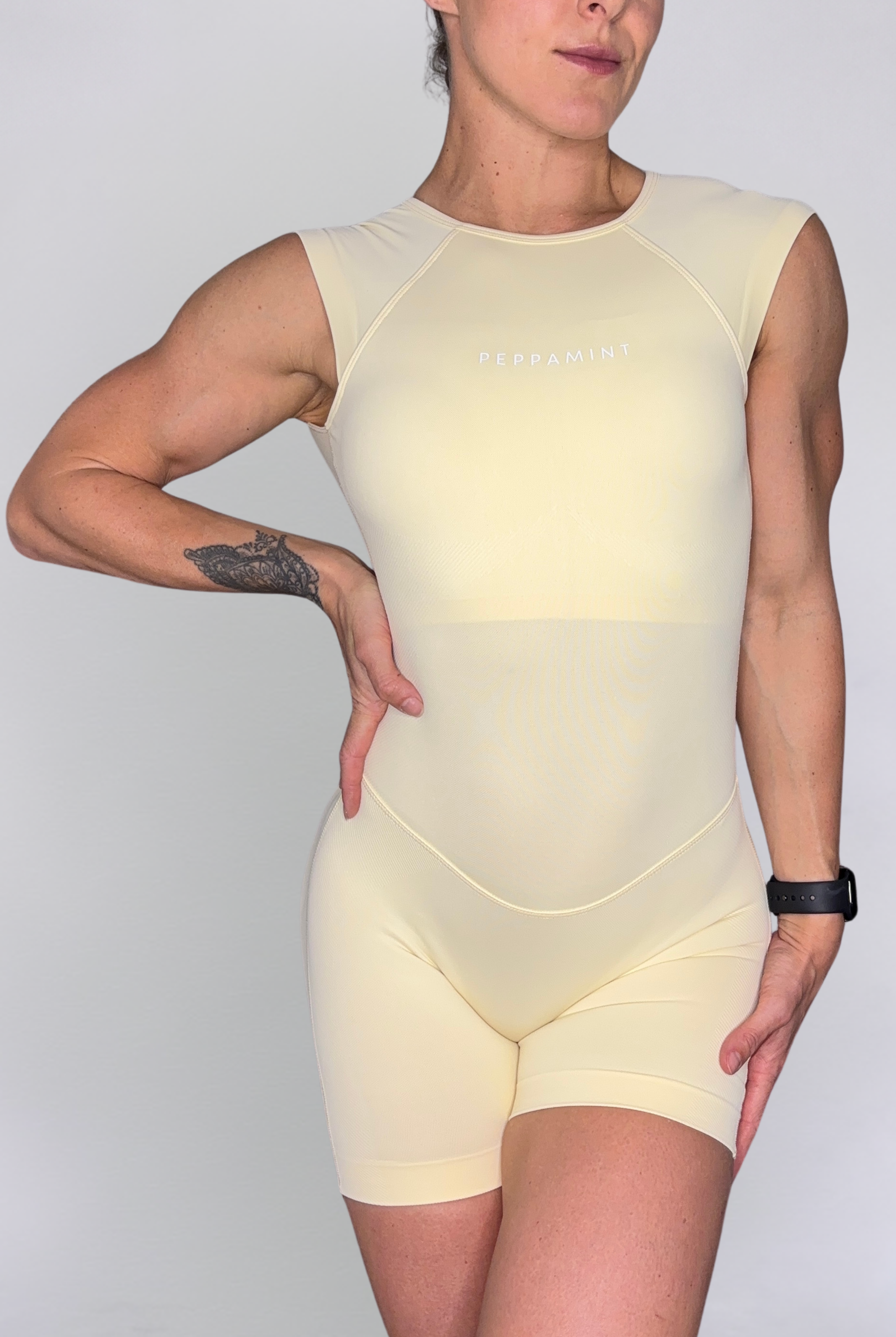A woman modelling a pale yellow sleeveless bodysuit with matching shorts against a light grey studio background. The bodysuit a small white 'PEPPAMINTI' logo on the centre chest.