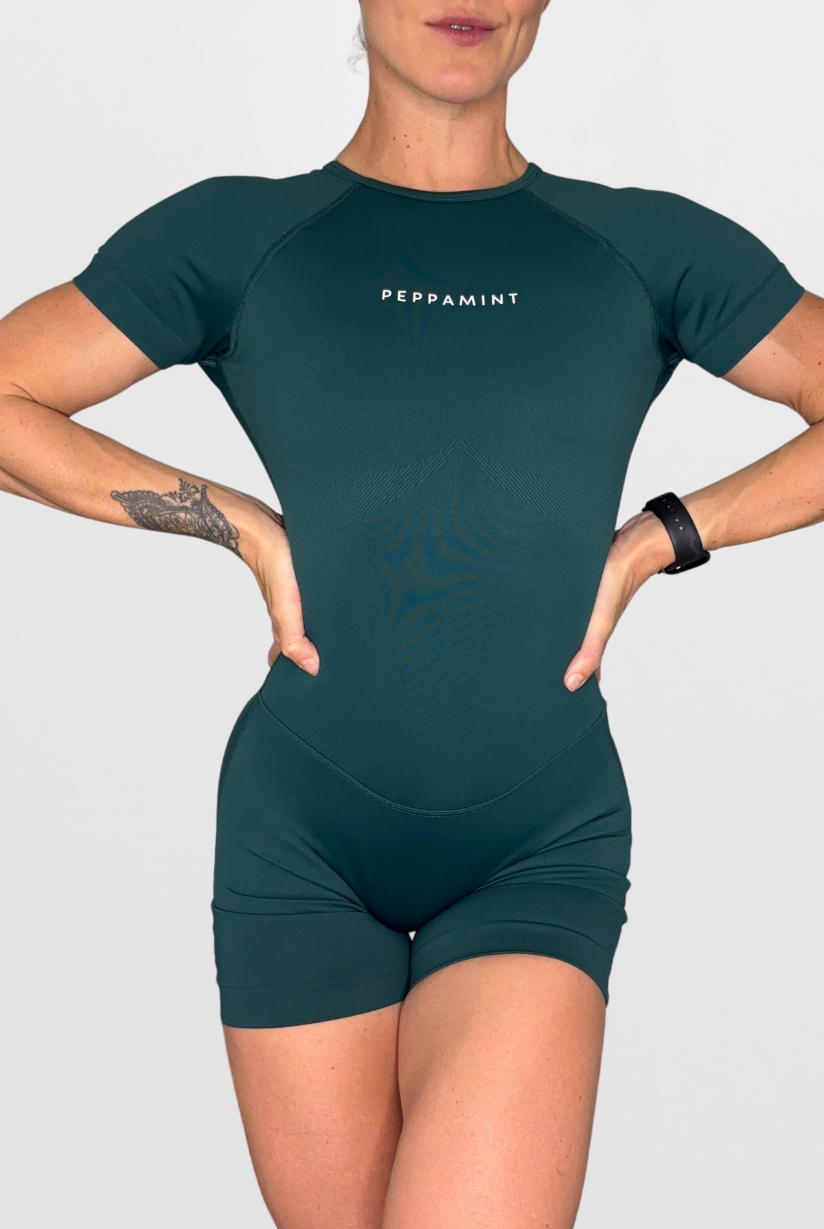 A woman modelling a dark Emerald Green short-sleeve athletic one-piece with 'PEPPAMINT' printed in white on the chest, set against a solid light grey background.