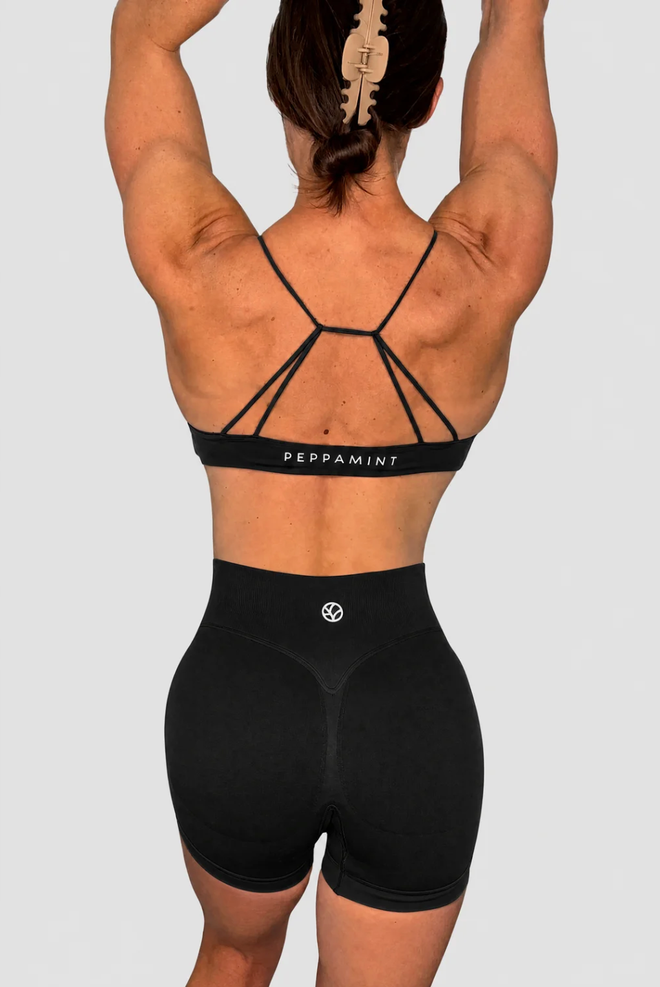 Rear view of a woman wearing a black strappy sports bra and matching high-waisted black athletic shorts against a light grey studio background.