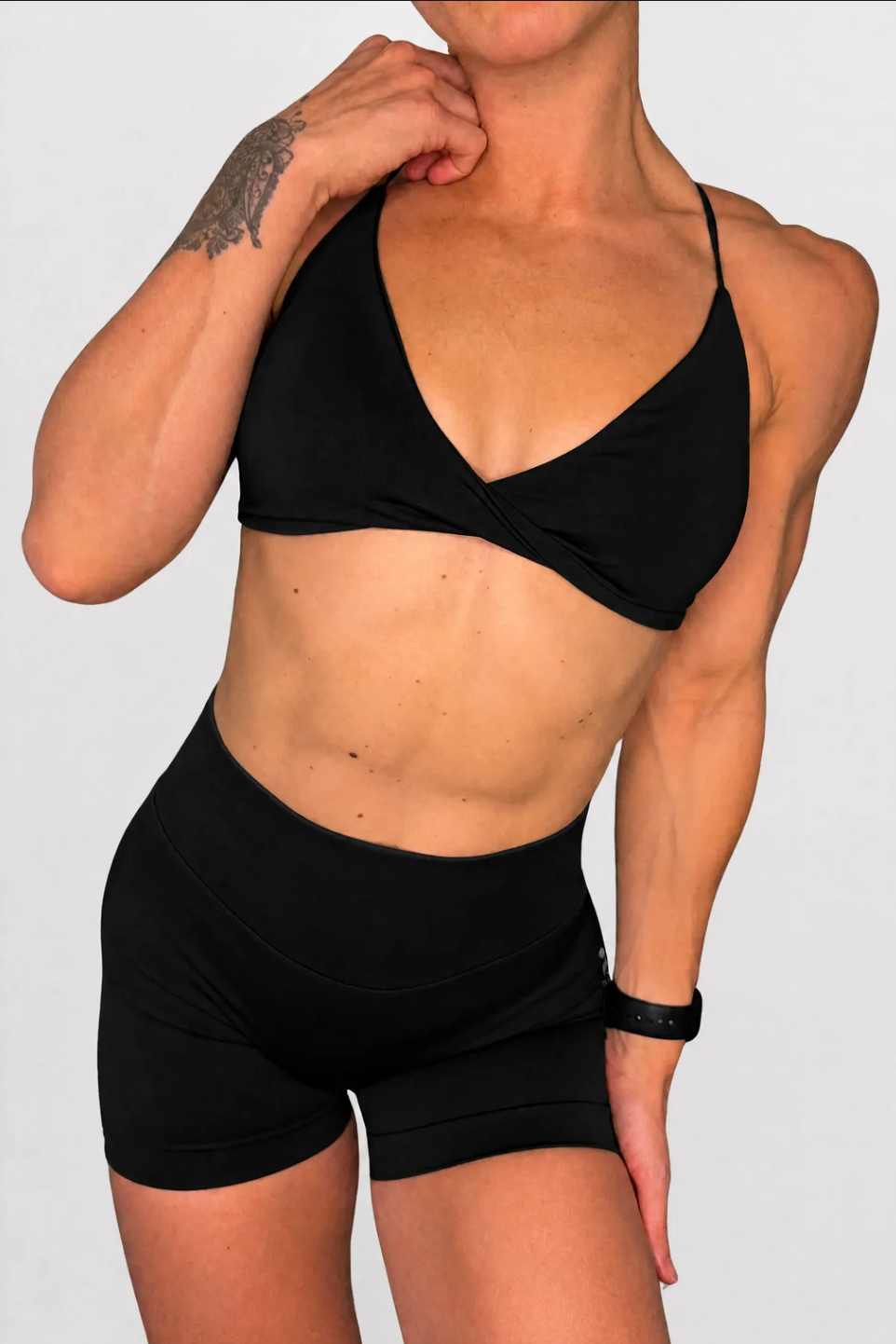 A woman modelling a black two-piece activewear set consisting of a twist style triangle sports bra and matching high-waisted shorts against a plain light grey background.