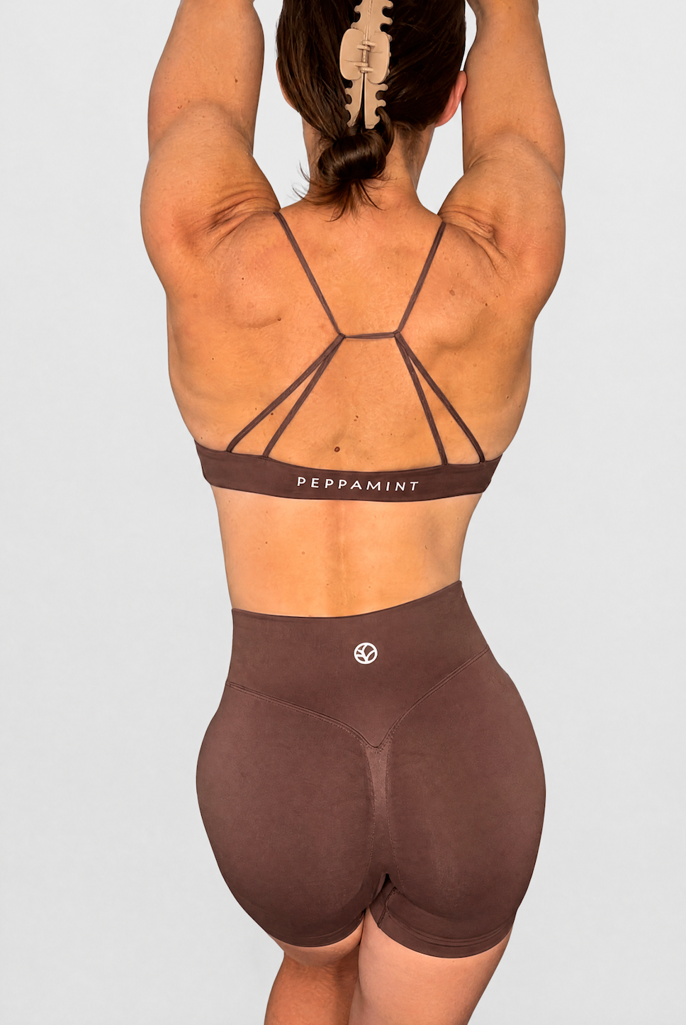 Rear view of a woman wearing a brown strappy sports bra and matching high-waisted shorts against a light grey studio background.