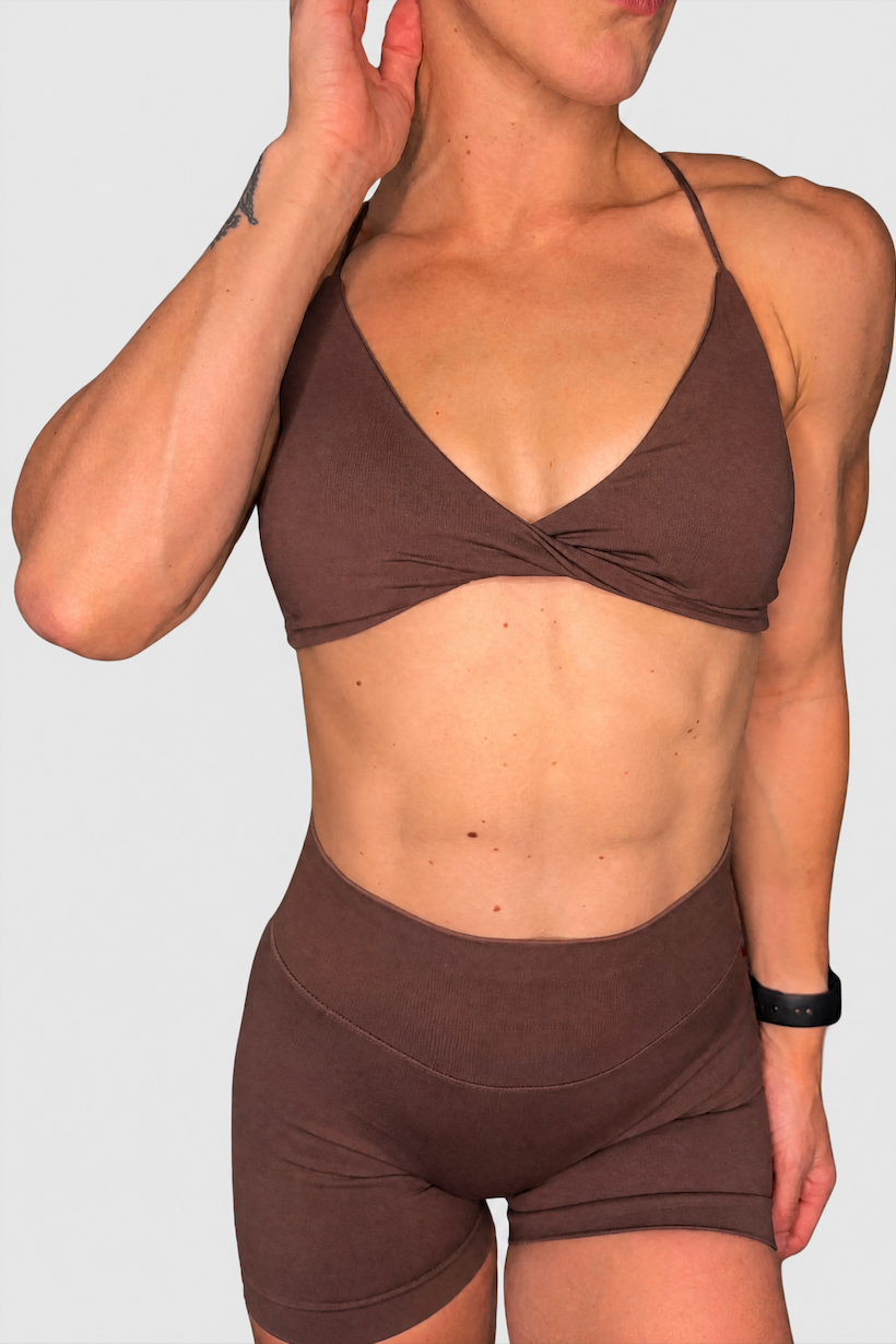 A woman modeling a matching brown activewear set consisting of a twisted crop top and high-waisted shorts, shown against a solid light grey background.