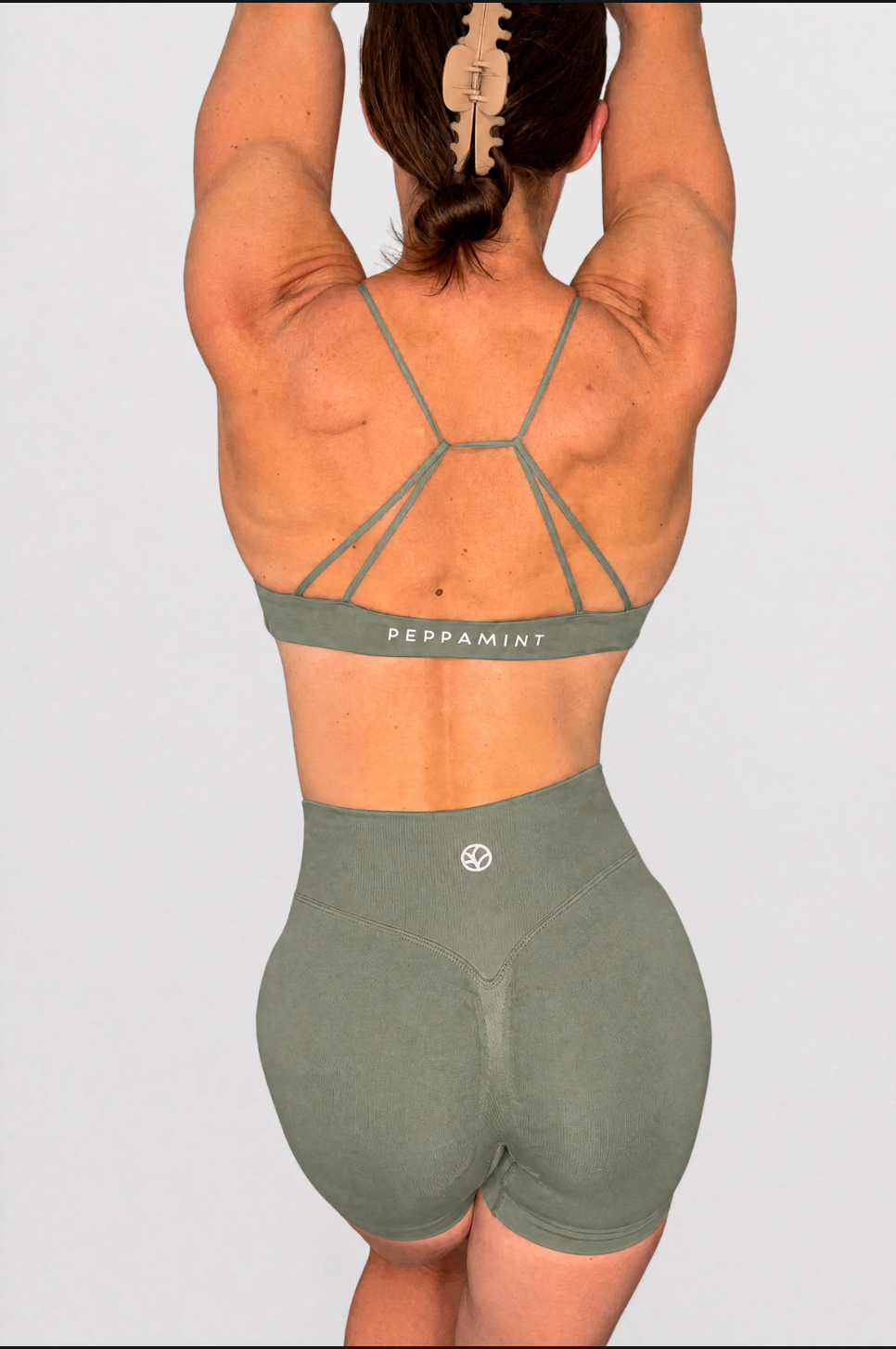 Rear view of a woman wearing an olive green athletic bra and matching high-waisted shorts against a light grey studio background.