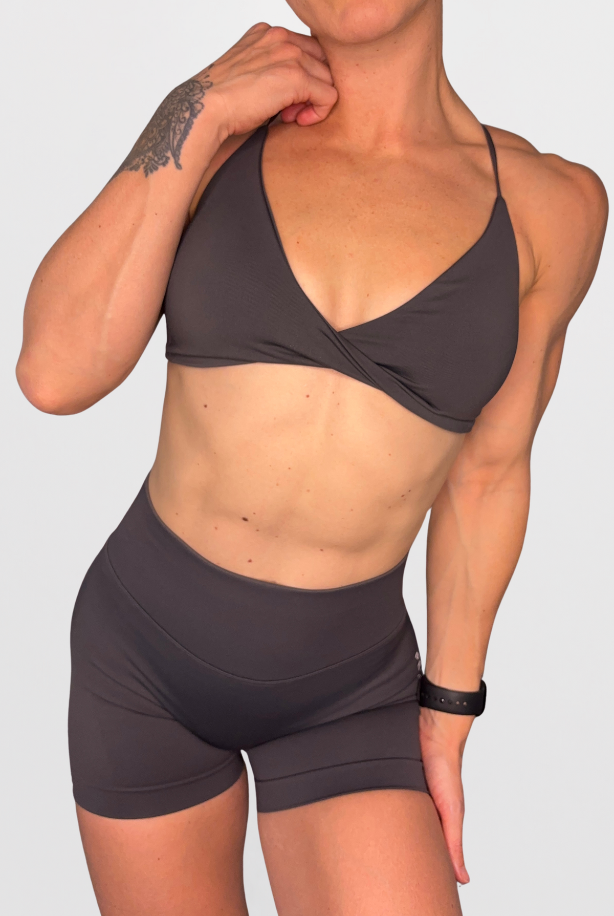 A woman modeling a dark grey athletic two-piece set consisting of a crossover sports bra and high-waisted shorts, shown against a solid light grey background.