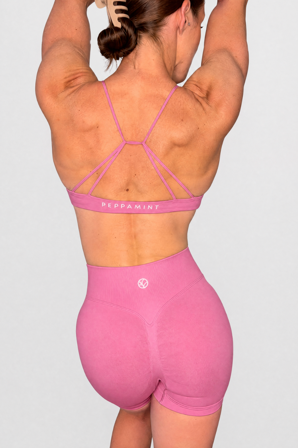 Rear view of a woman wearing a rose pink strappy sports bra and matching high-waisted biker shorts against a light grey studio background.
