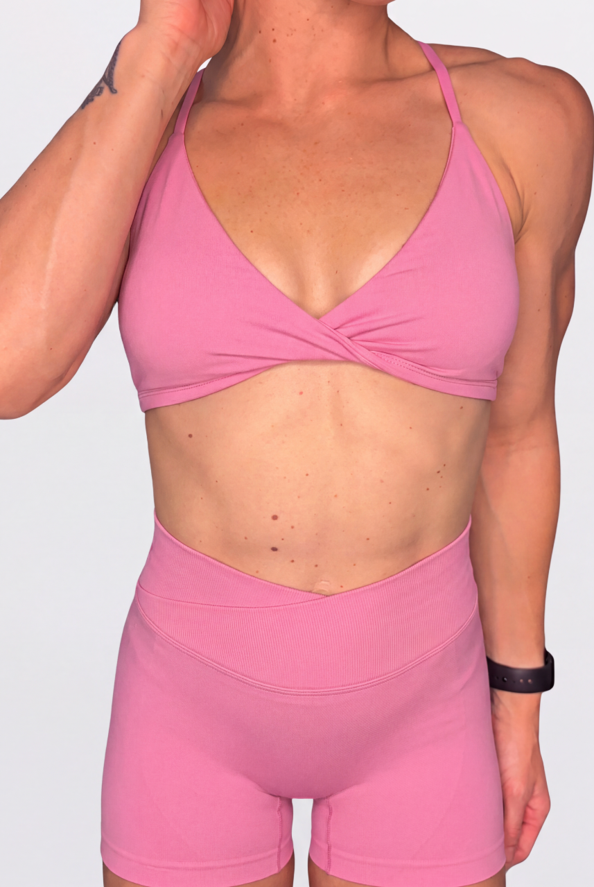 A woman wearing a rose pink sports bra with a twisted front detail and matching high-waisted pink athletic shorts, shown against a light geay studio background.