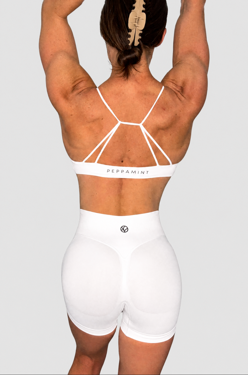 Rear view of a woman wearing white high-waisted athletic shorts and a matching white sports bra with strappy back detailing, on a light grey background.