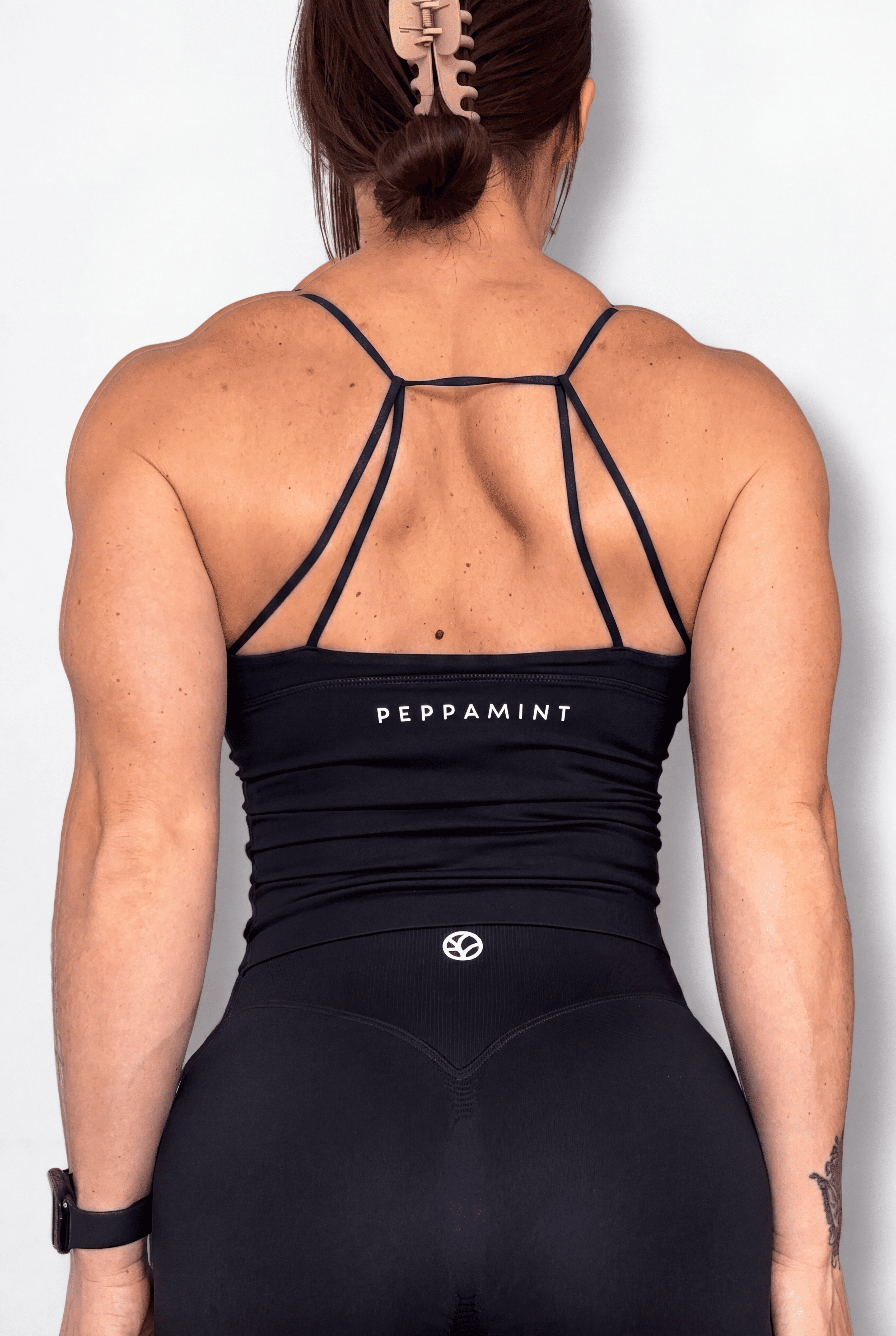 Rear view of a woman wearing a black athletic tank top and matching leggings. The top has thin crisscross shoulder straps and the word 'PEPPAMINT' printed in white across the upper back.