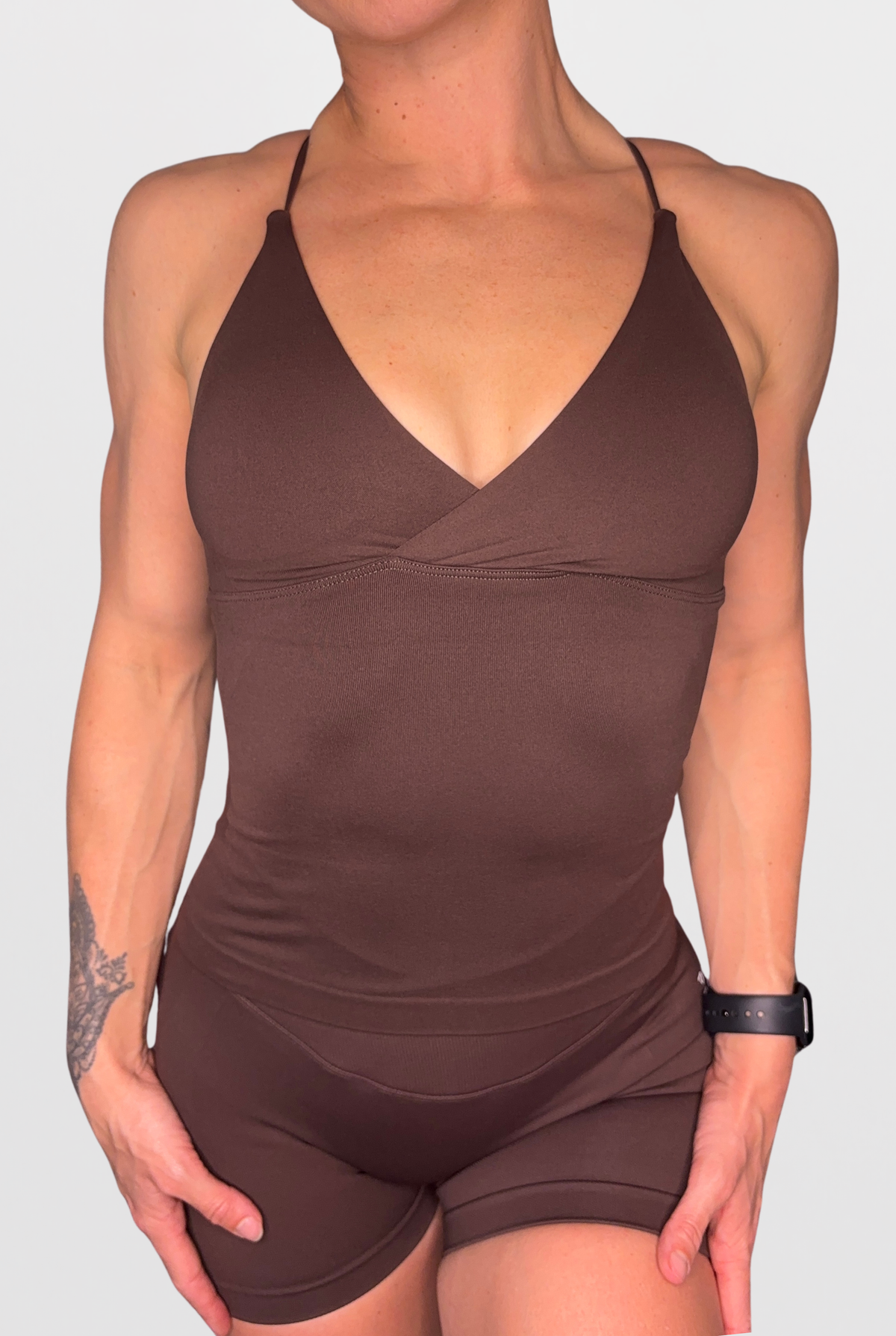 A woman wearing a dark brown athletic tank top with a wrap-style V-neck bodice and matching fitted shorts, shown against a light grey studio background.