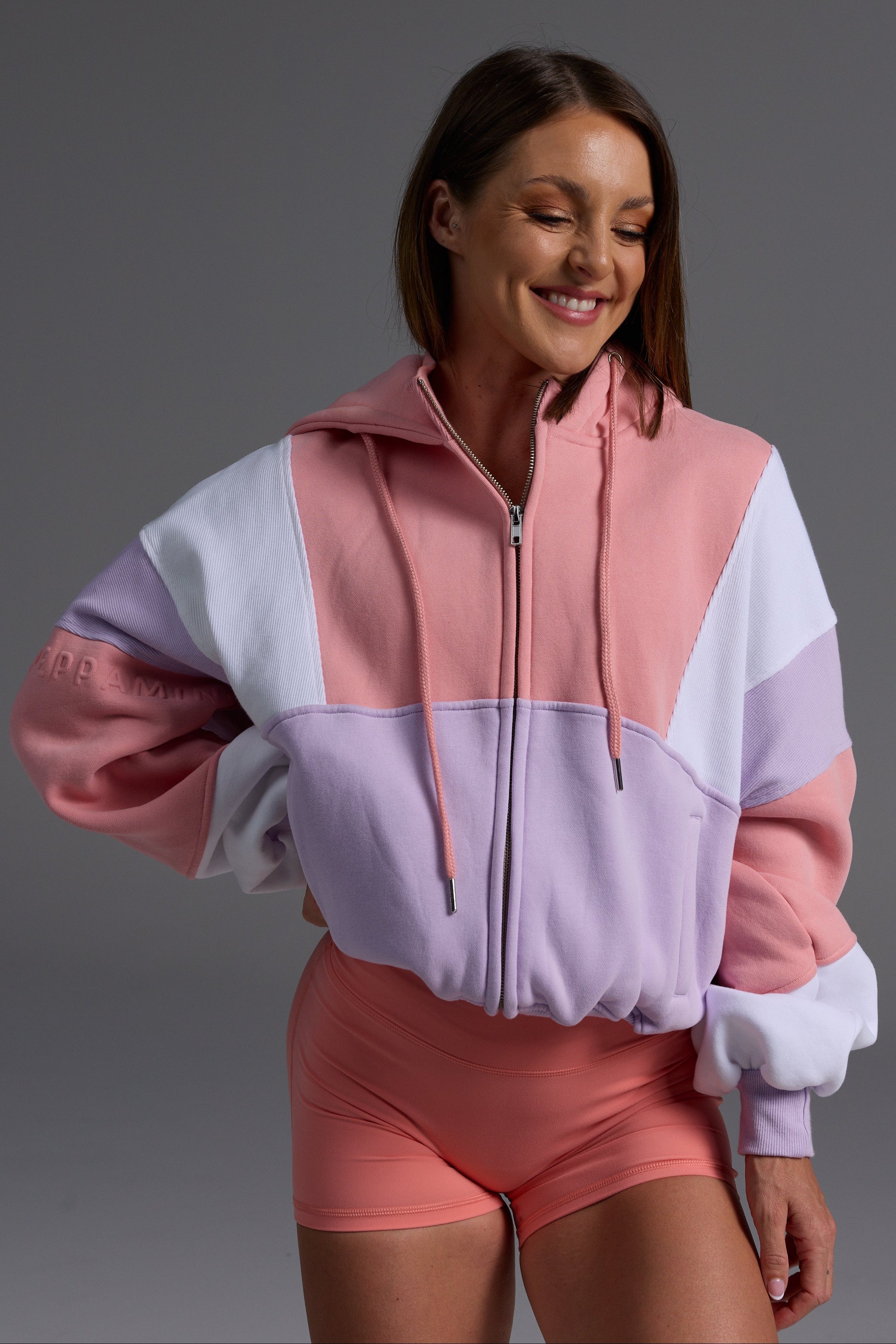 Woman in peach, lilac and white color-blocked oversized cropped fleece hoodie with matching coral bike shorts, smiling against grey background