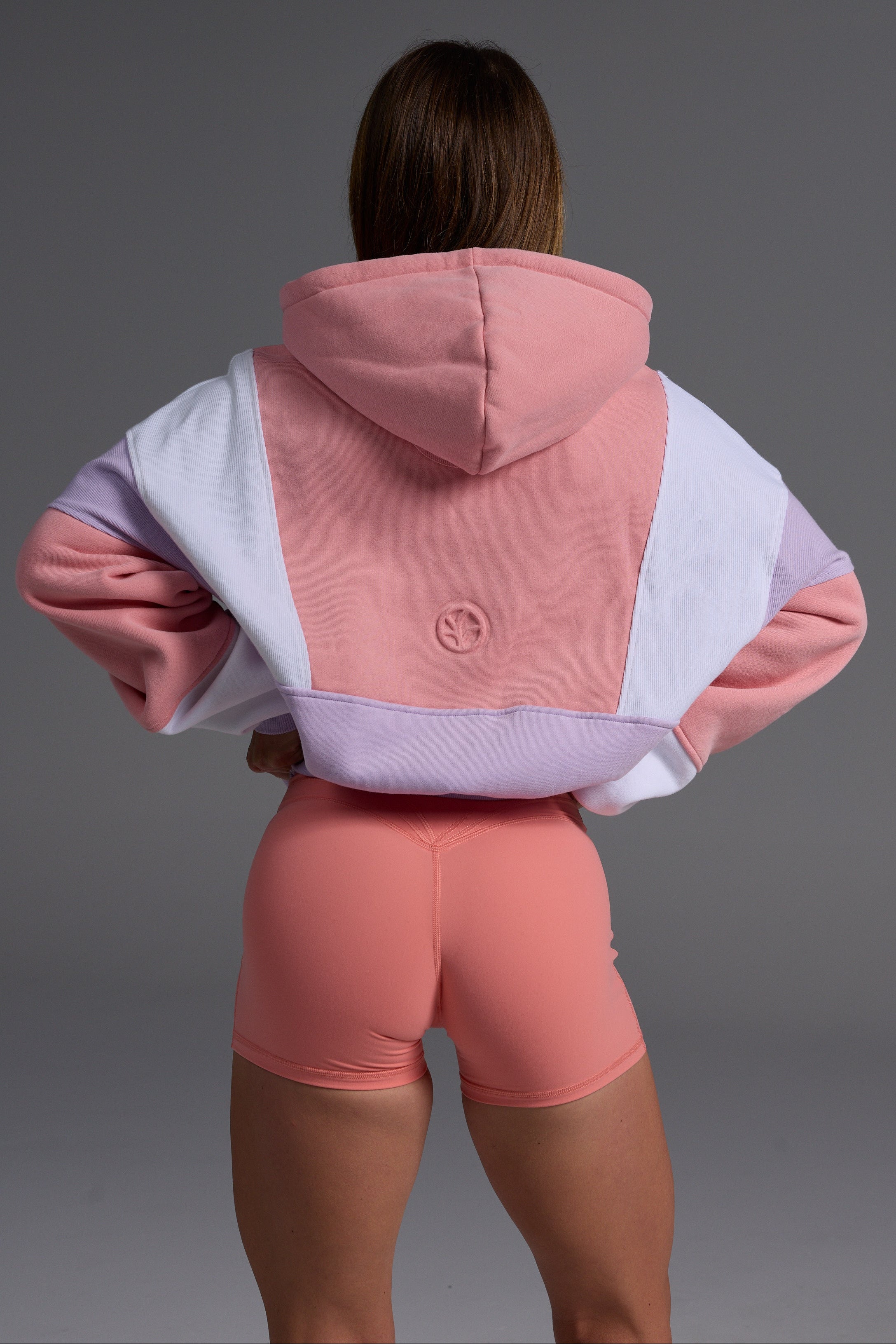 Woman in peach, lilac and white oversized fleece hoodie with coral bike shorts in confident pose against grey background 