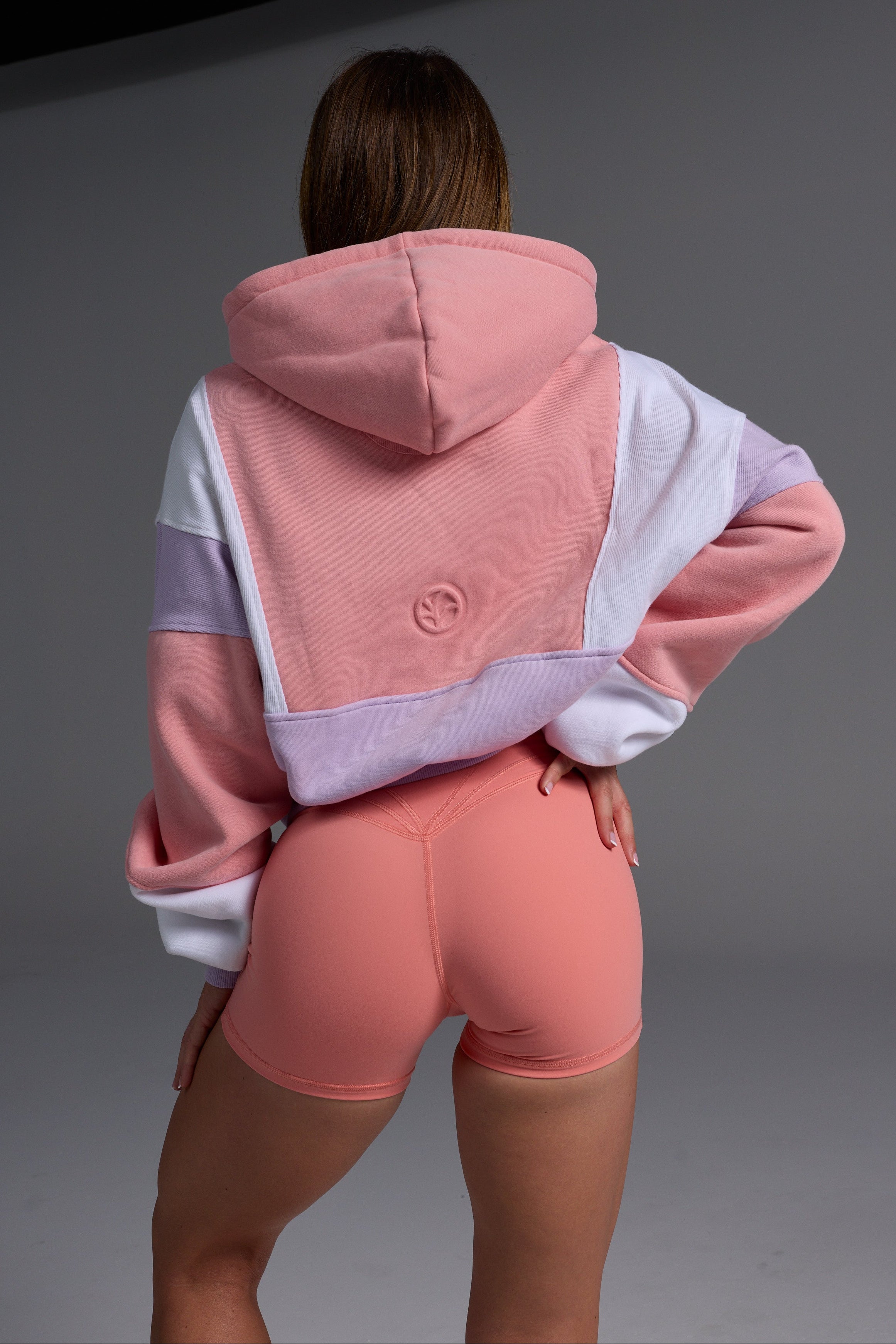 Back view of woman in pink peach, lilac and white colour-blocked fleece hoodie with coral bike shorts in studio setting